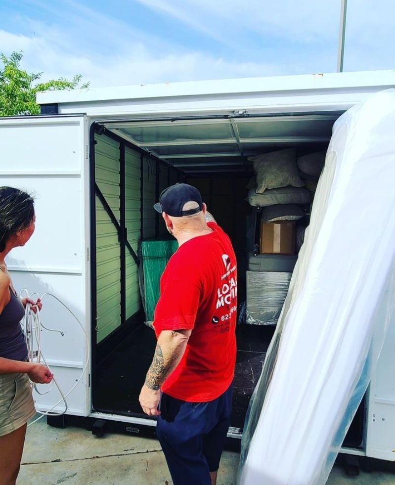 Two people load belongings into a moving truck. The man in red points inside. A mattress leans against the truck.
