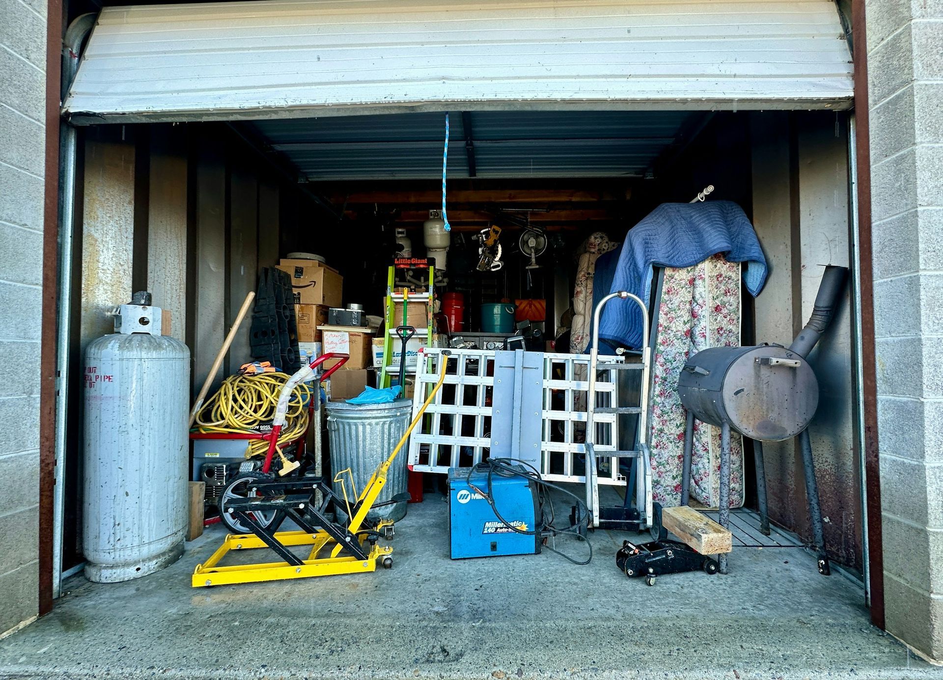 Storage unit filled with items: propane tank, boxes, tools, metal smoker.