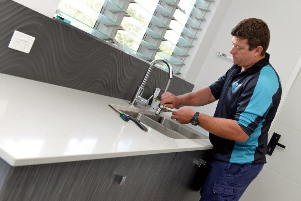 Plumber fixing a faucet in a modern bathroom. He wears a blue shirt, working on a white countertop — Top To Bottom Plumbing in Garbutt, QLD