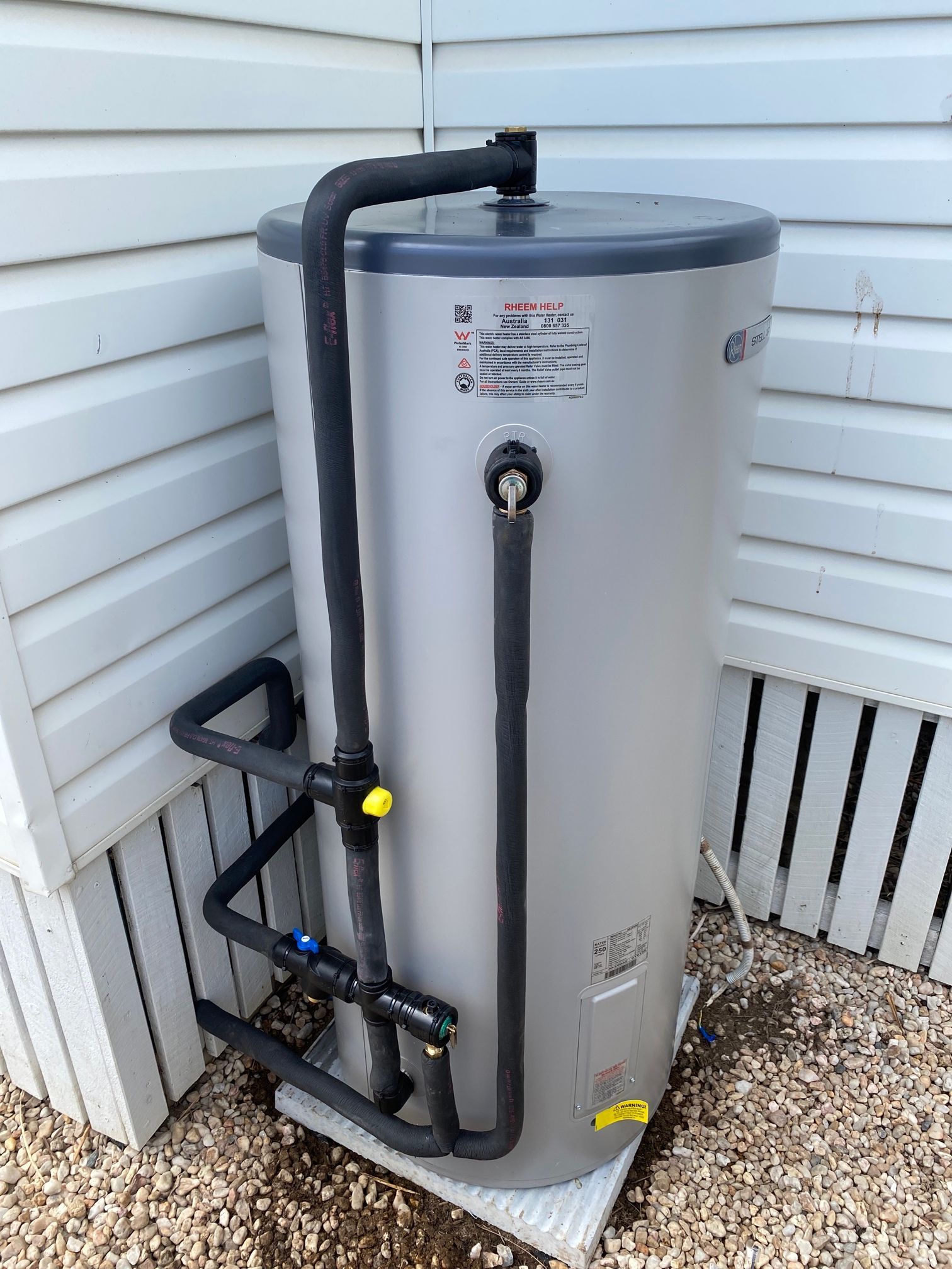 Silver water heater tank with black pipes and a yellow valve, outdoors — Top To Bottom Plumbing in Garbutt, QLD