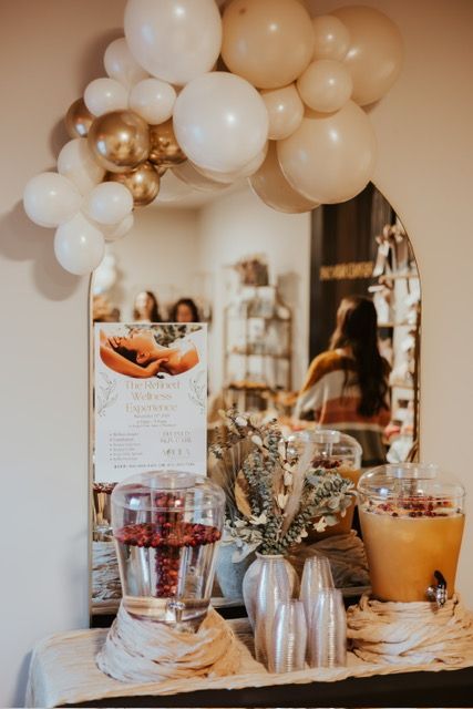 A party drink station with two dispensers and a balloon arch over a mirror, featuring white, gold, and tan decor.