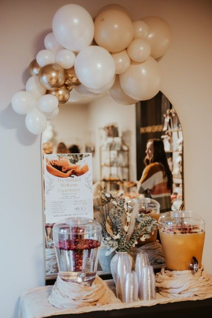 A beverage station in front of an arched mirror features a balloon arch, two glass dispensers, and a menu sign.
