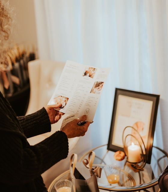 A person in a dark sweater reads a pamphlet near a small table holding a framed print, a lit candle, and a glass of juice.