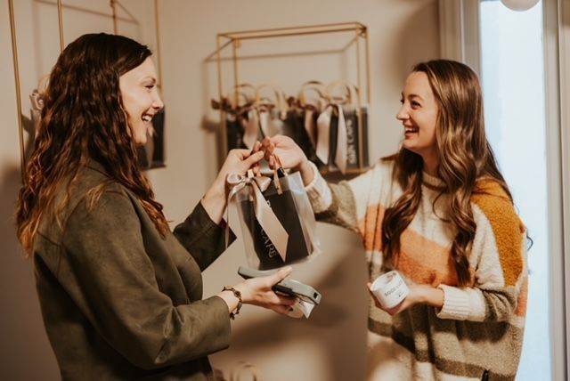Two people smiling while exchanging a gift in a well-lit indoor space with decorative shelves in the background.