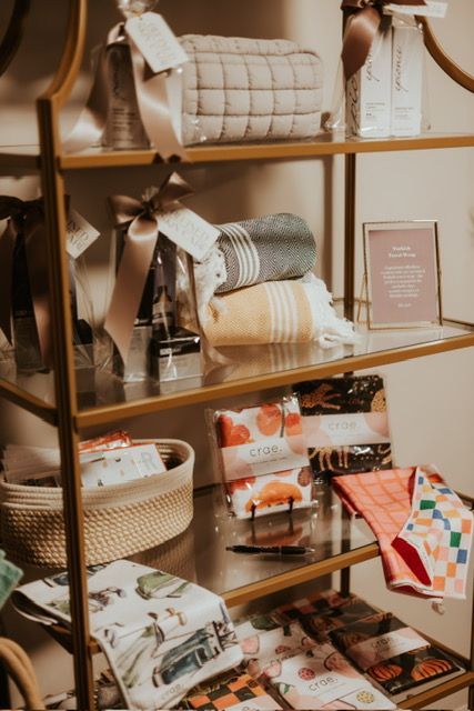 A gold-toned metal shelving unit displays home goods, including textiles, wrapped gift sets, and a woven storage basket.