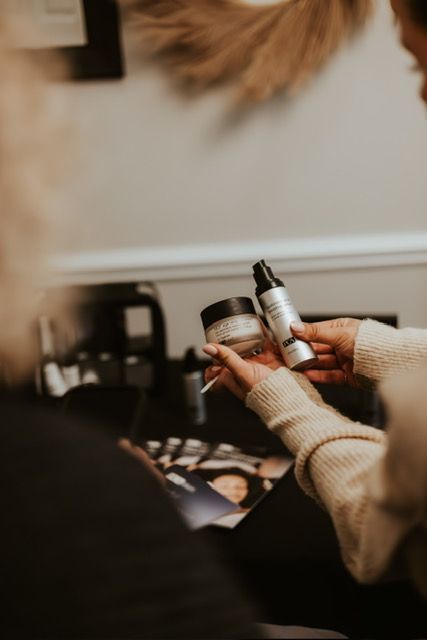 A person wearing a beige sweater holds a small jar and a bottle of skincare products in a softly lit, modern setting.