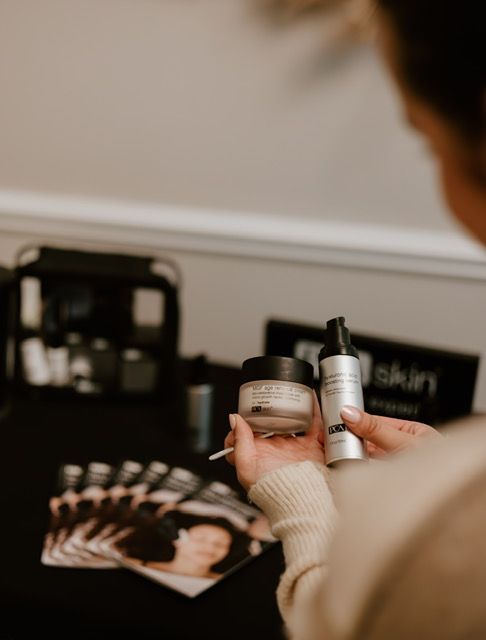 Hands hold a skincare jar and pump bottle in front of a promotional display and brochures on a table.