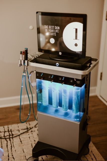 A HydraFacial machine on a rolling cart with a display screen, treatment wands, and four illuminated serum bottles.