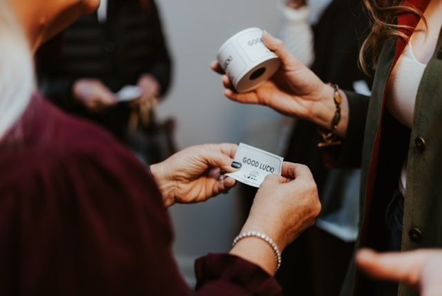 Two people exchange a small white raffle ticket while one holds a roll of tickets in an indoor setting.