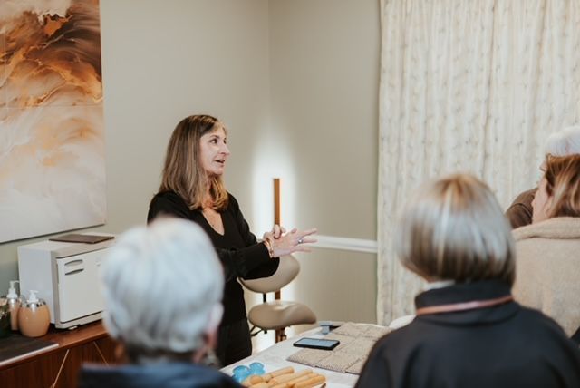 A speaker gestures while talking to a small group in a room with a painting, a white towel warmer, and a tall lamp.