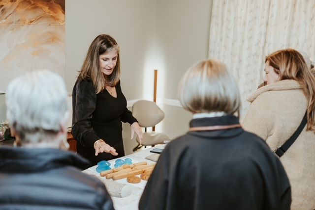 A woman gestures toward wooden blocks and blue objects on a table while speaking to a small group in a bright room.