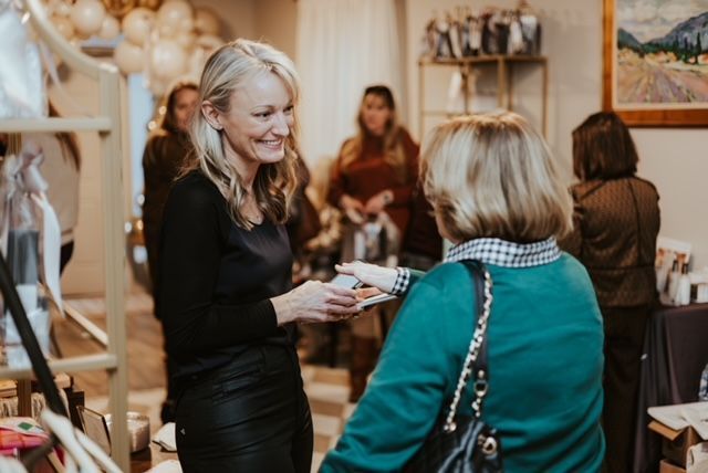 A woman in a black shirt smiles while handing a small object to another person in a teal sweater at an indoor event.