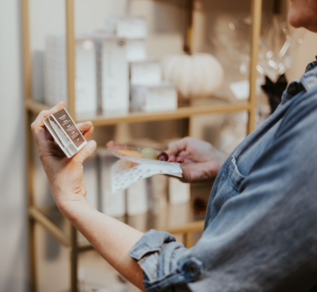 A person in a denim shirt examines a small, brown product box while holding a napkin with a single grape in a store.