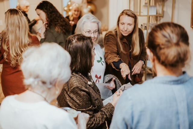 People gather in a warmly lit room, reviewing documents and conversing while some examine items on a display table.