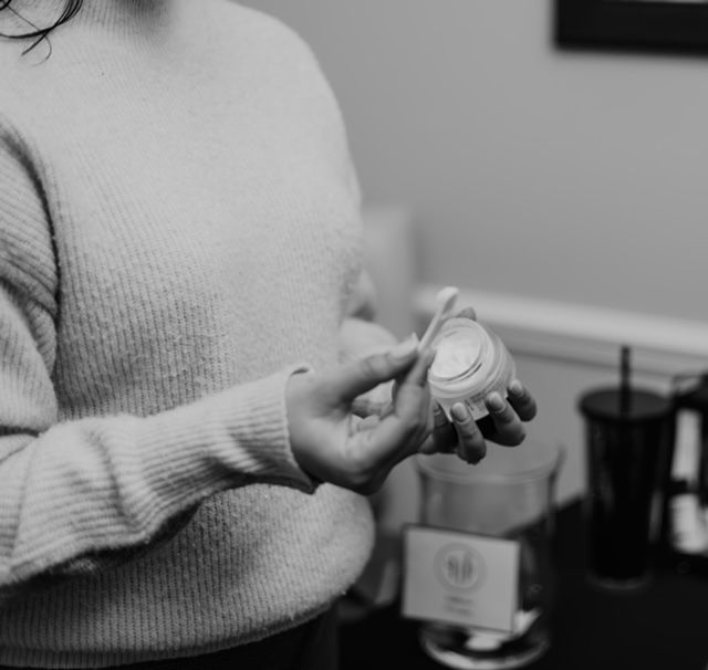 A person holds a small jar of cream and a spatula, preparing to apply a skincare product in a professional setting.