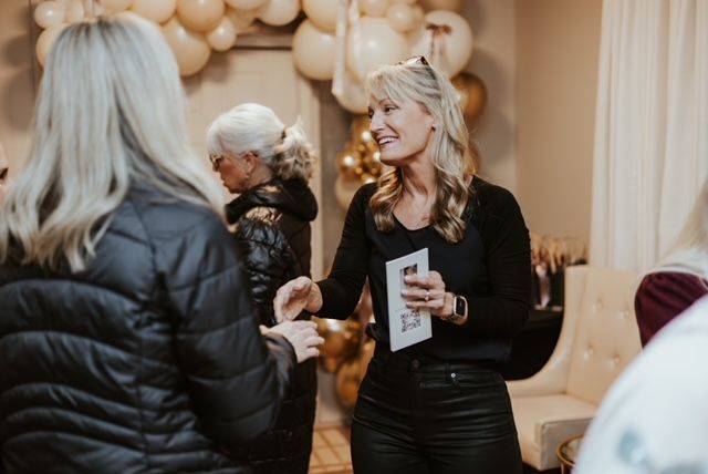 Three people in casual attire converse at an indoor social gathering decorated with beige and gold balloons.