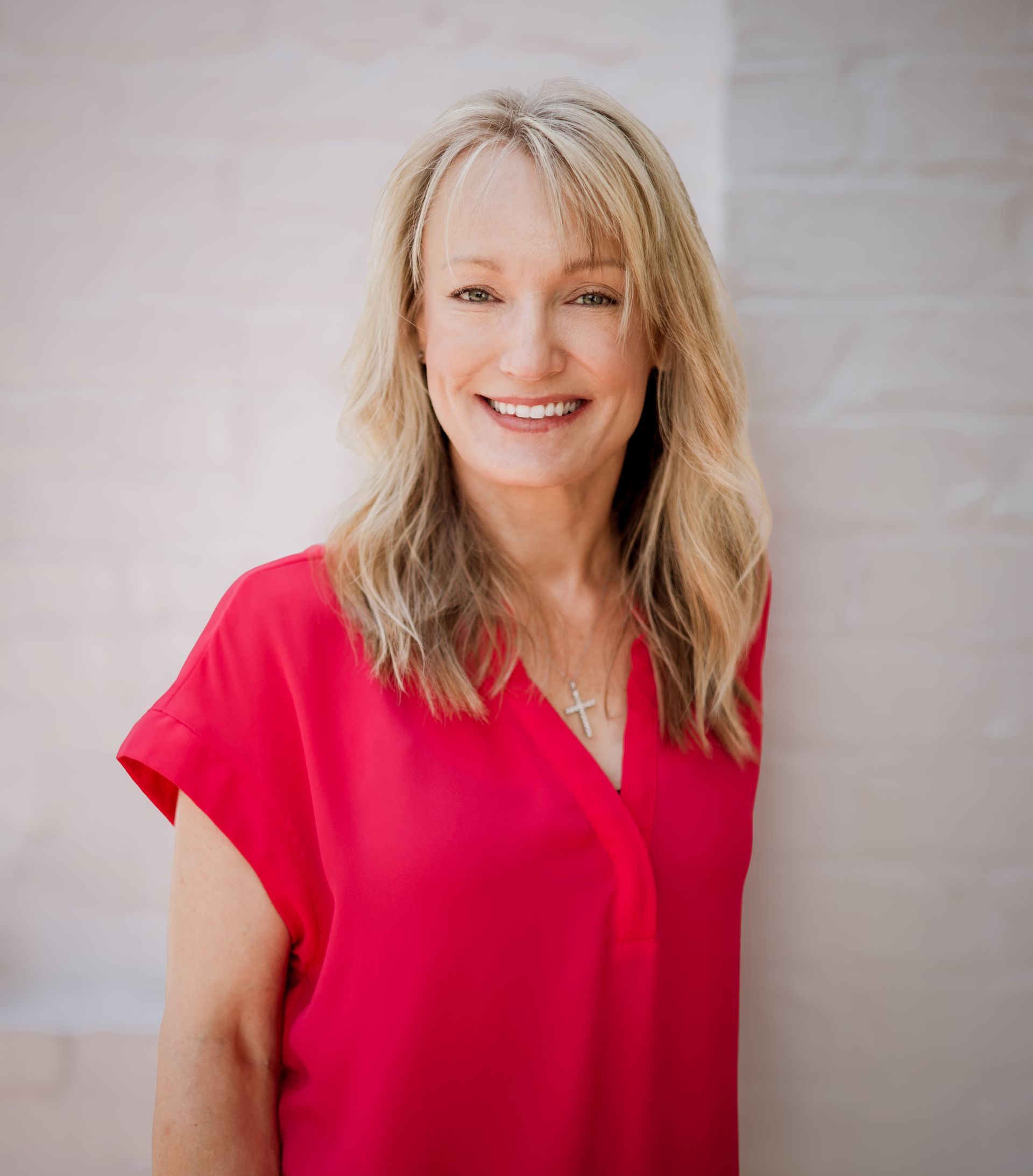 A woman in a red shirt is smiling and standing in front of a white wall.
