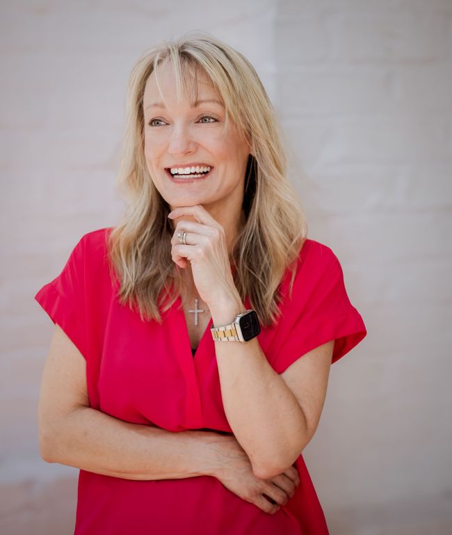 A woman in a red dress is smiling and holding her hand to her chin.