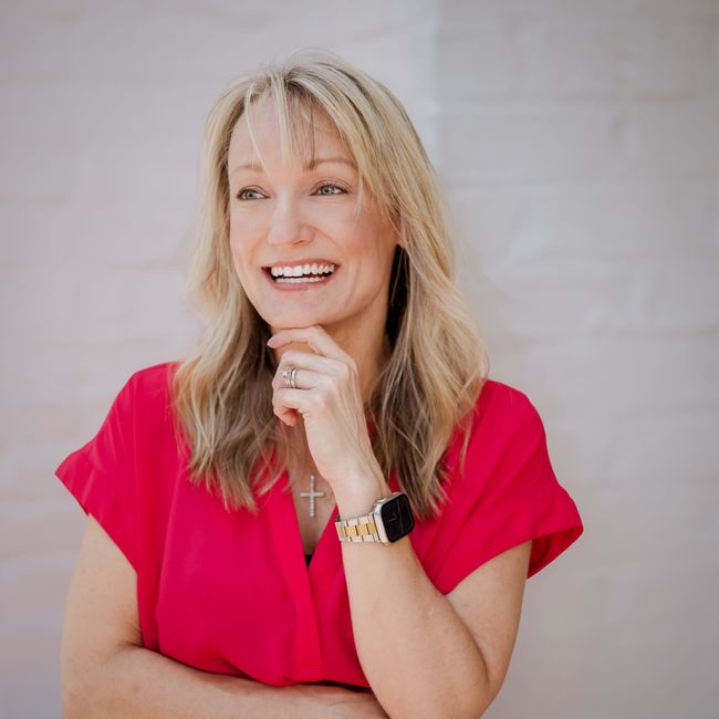A woman in a red dress is smiling and holding her hand to her chin.