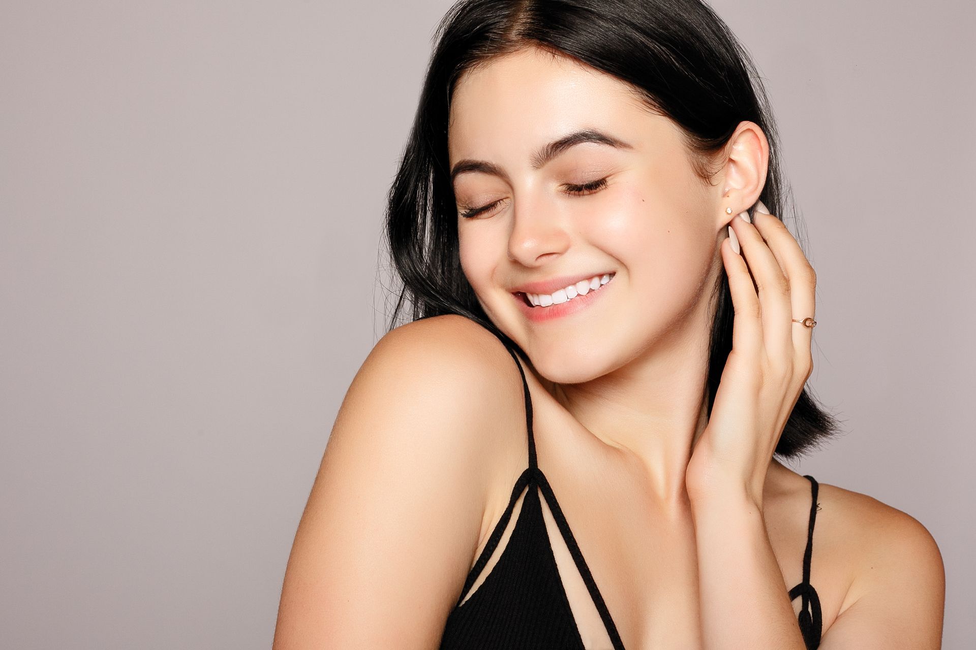A woman in a black dress is smiling with her eyes closed and touching her ear.