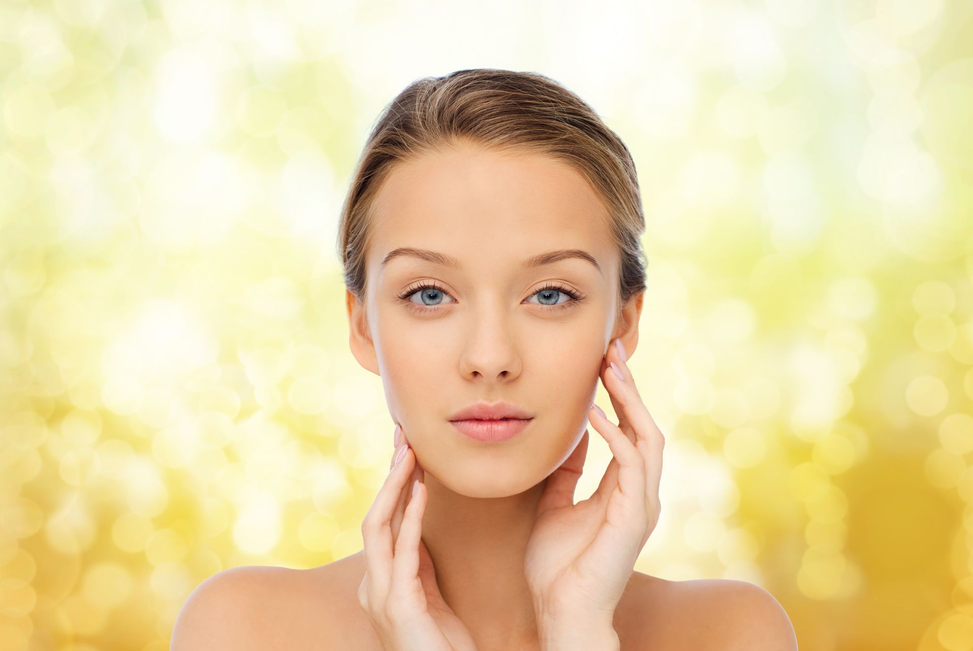 Woman with fair skin, gently touching her face, against a blurry, yellow background.