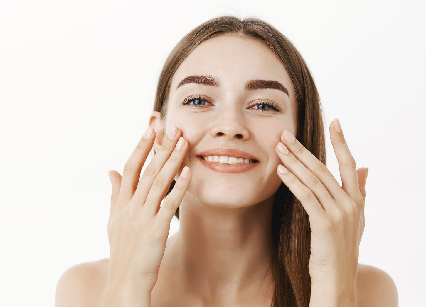 A smiling person with long brown hair gently touches their cheeks with both hands against a plain white background.
