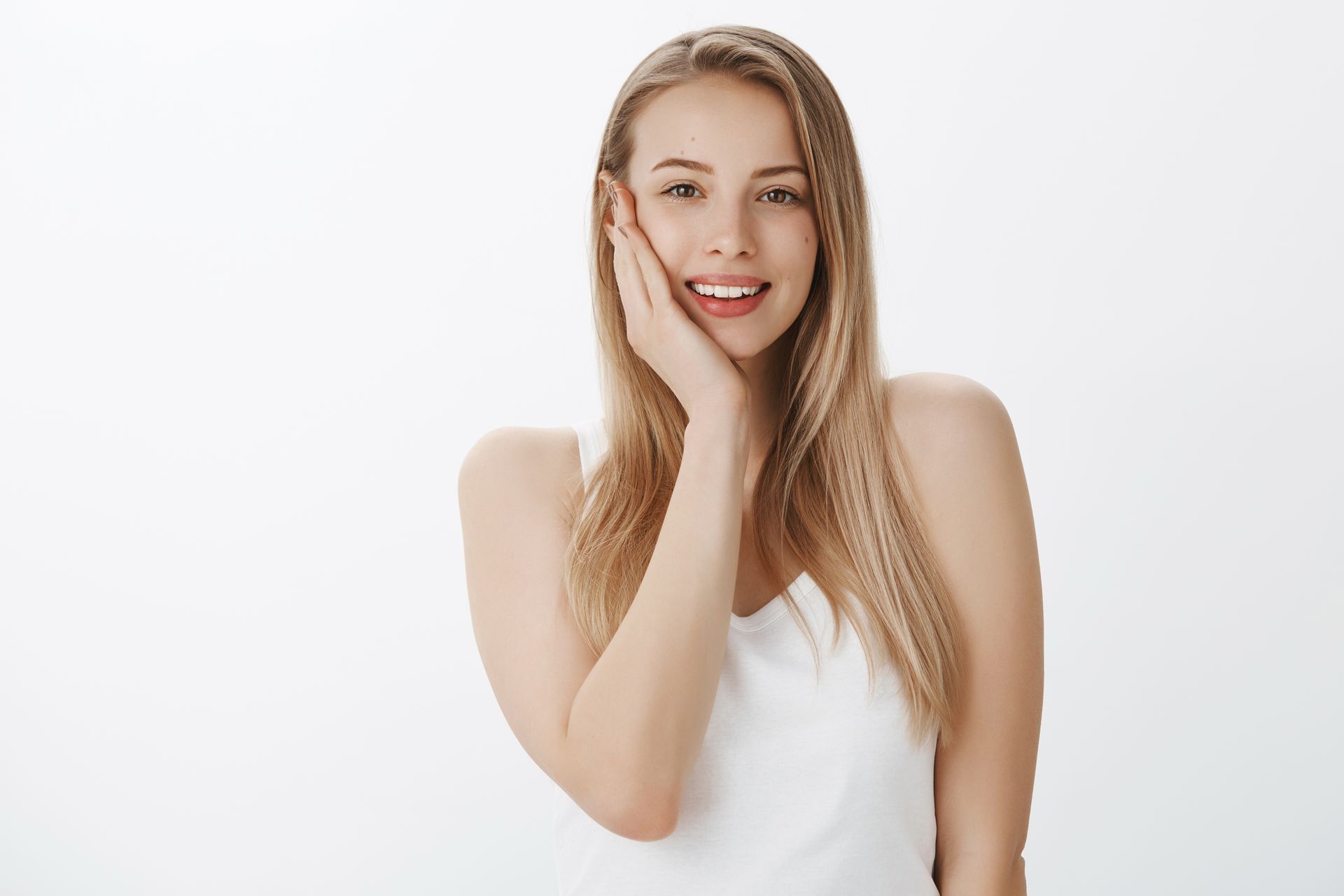 A woman in a white tank top is smiling and touching her face.