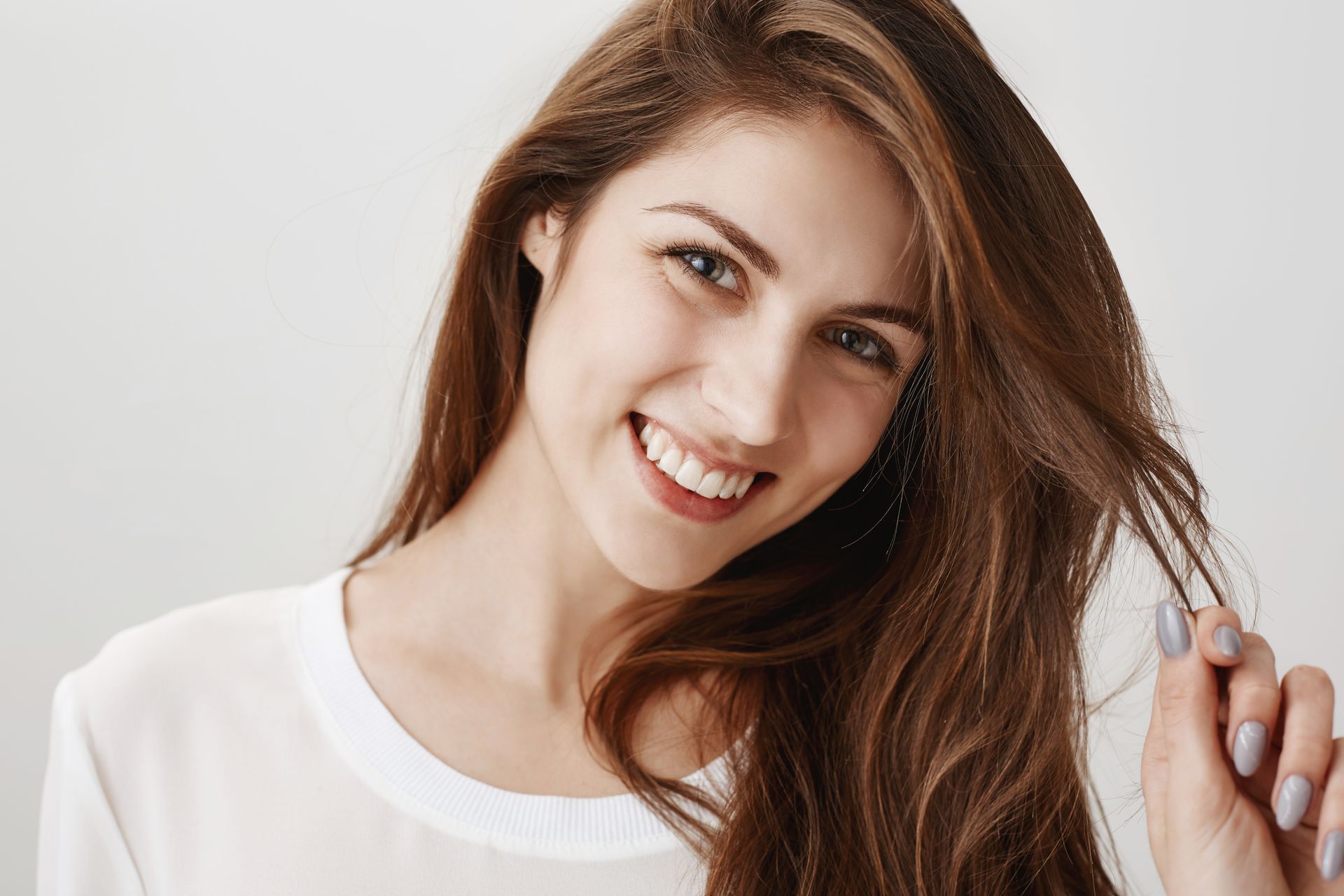 A smiling person with long brown hair, wearing a white shirt against a light background, holds a strand of their hair.