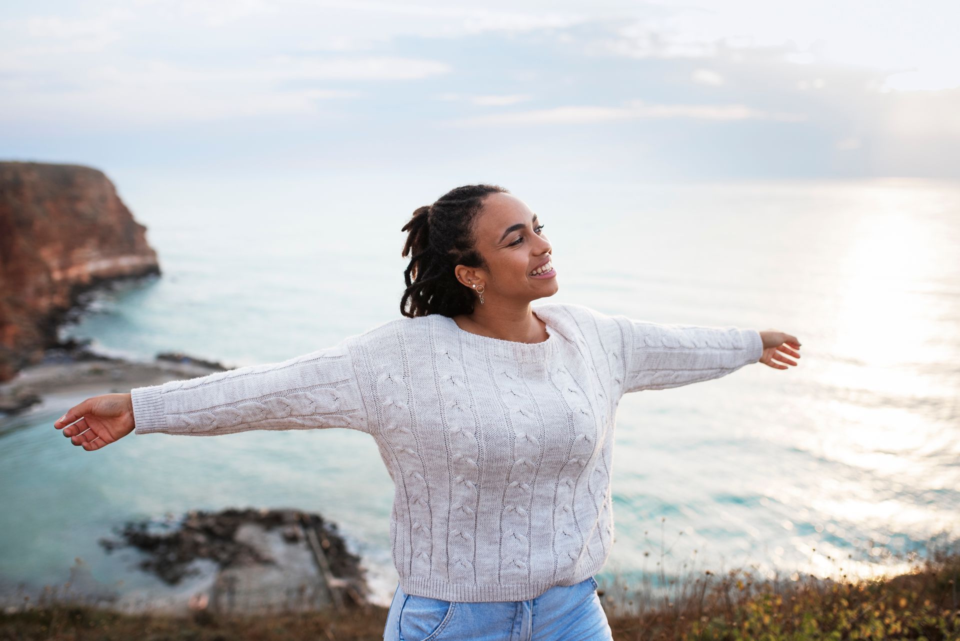 A woman is standing on a cliff overlooking the ocean with her arms outstretched.