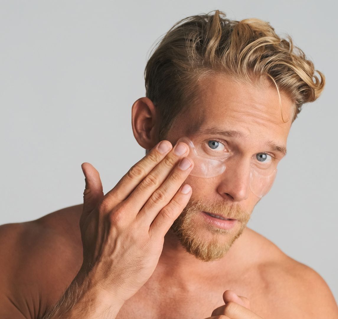 A shirtless man is applying lotion to his face.