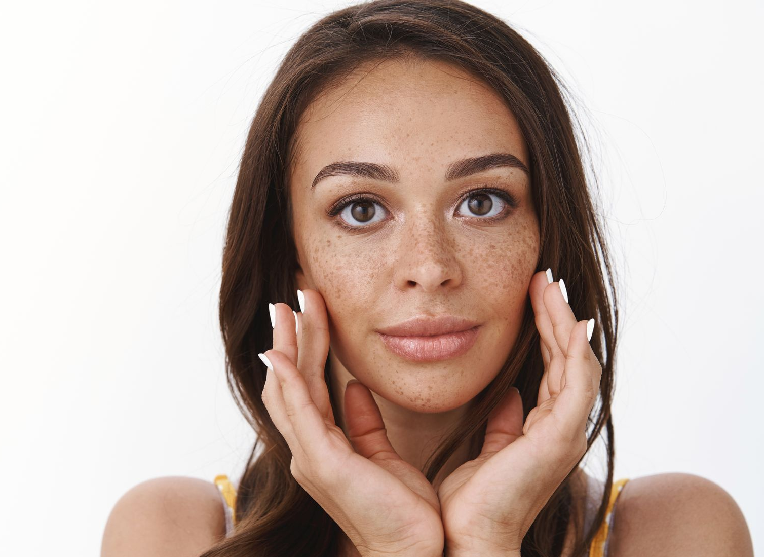 A person with freckles looking directly at the camera, holding both hands gently against their face against a white backdrop.