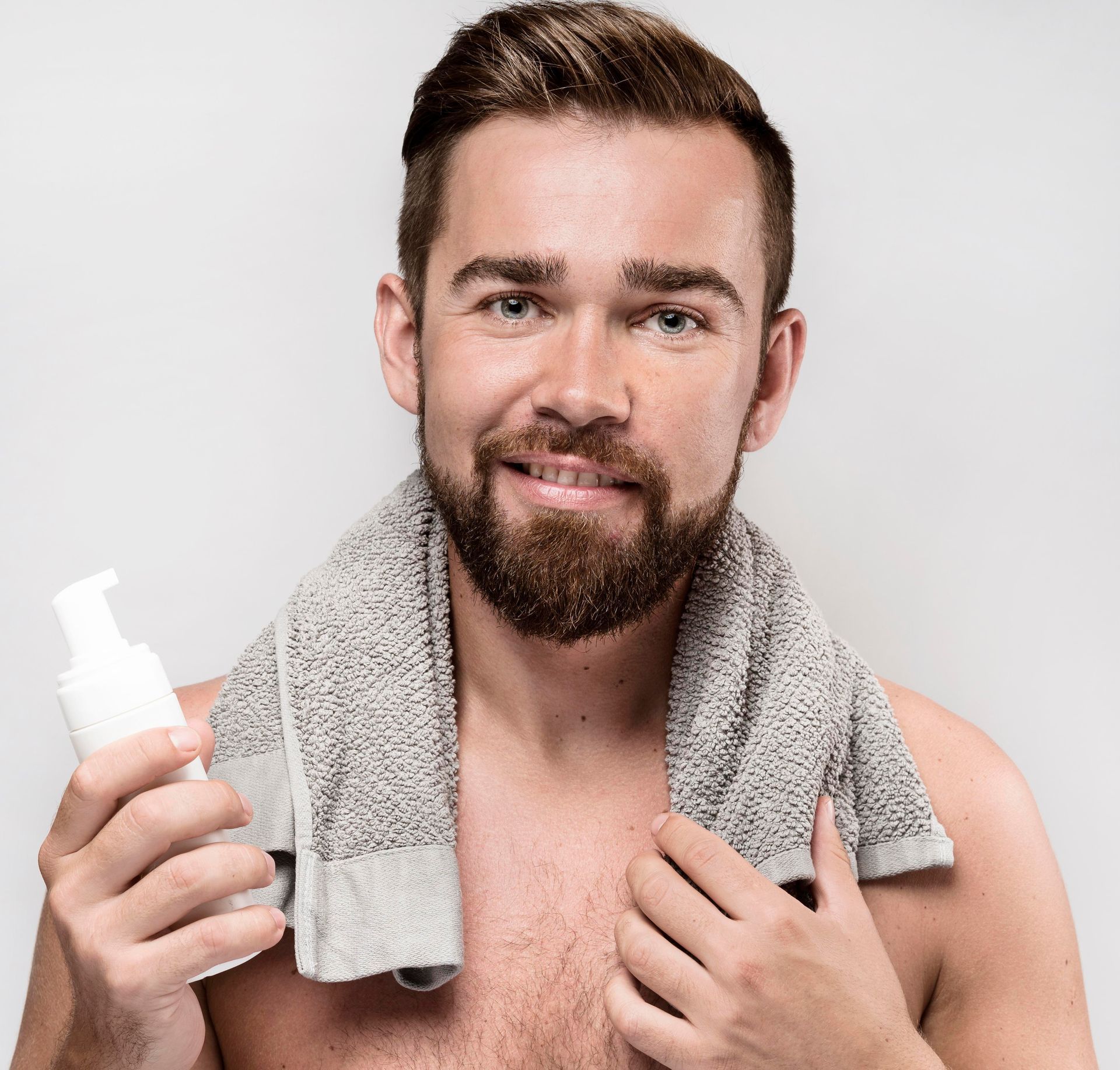 A shirtless man with a towel around his neck is holding a bottle of lotion.