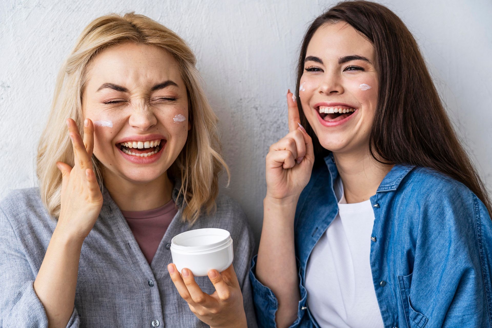 Two women are laughing while applying cream to their faces.