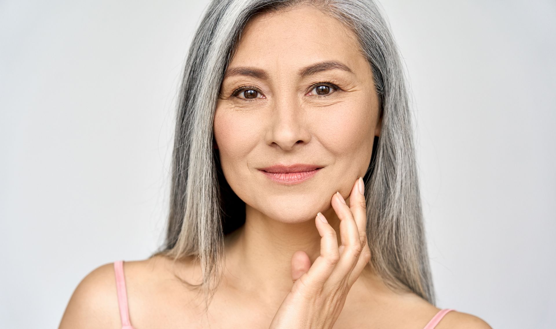 A smiling person with long gray hair touching their cheek against a neutral background.