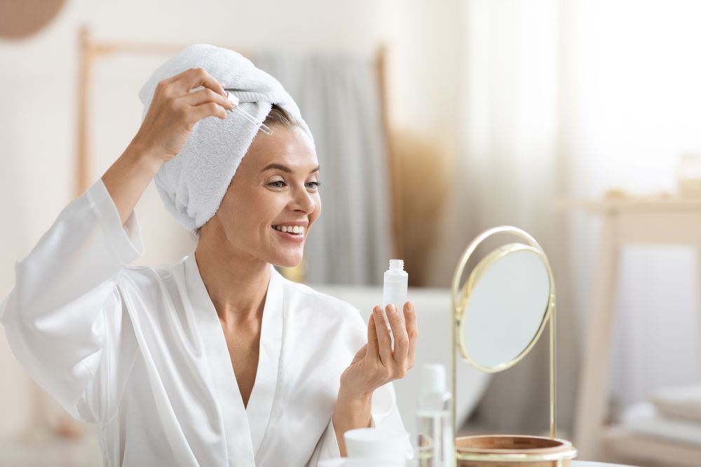 A smiling person with a white towel wrapped around their head holds a small cosmetic bottle while looking in a mirror.