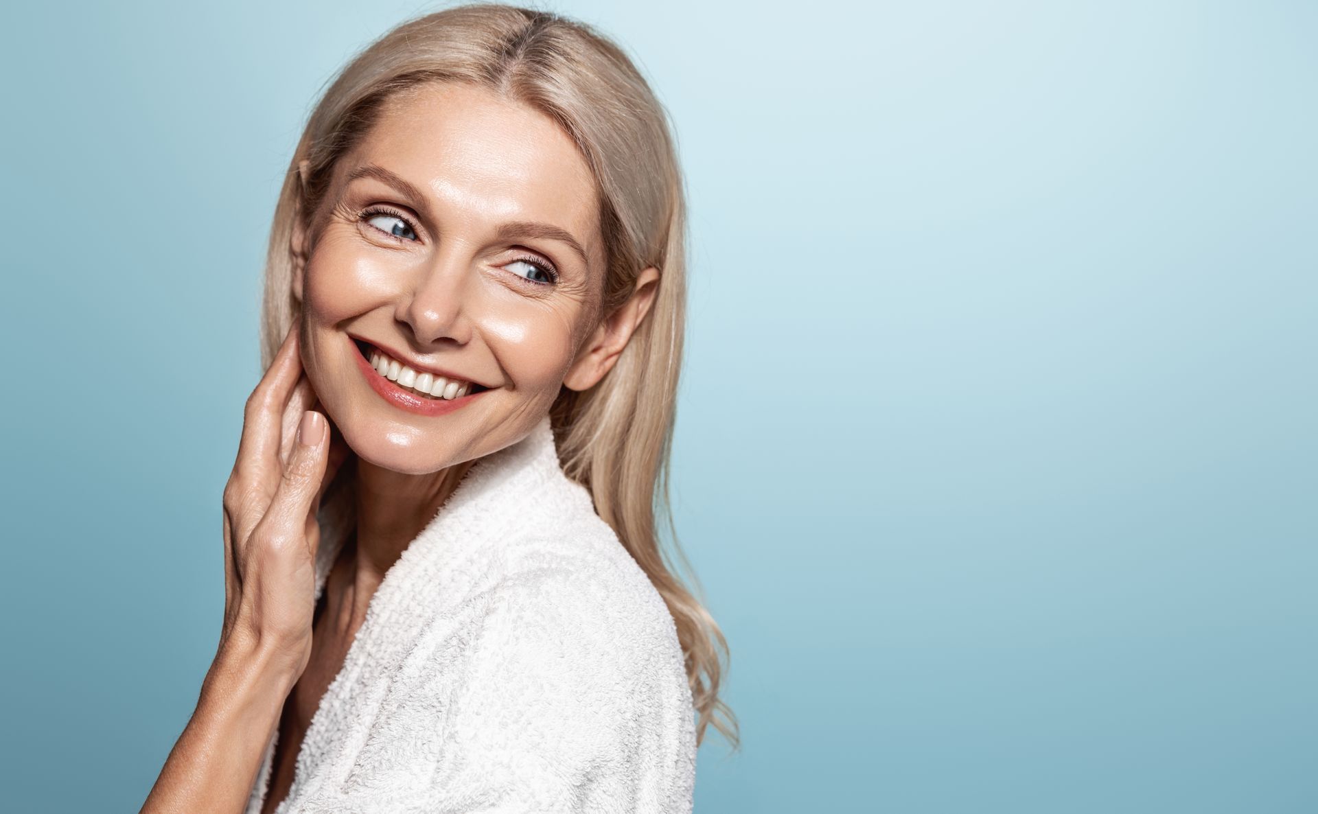 Woman in white robe smiles, touching her neck, with blonde hair, against a light blue background.