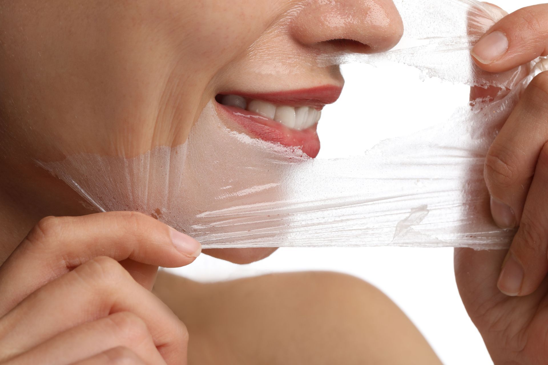 Woman peeling a facial mask from her face, smiling with her mouth open, against a white background.