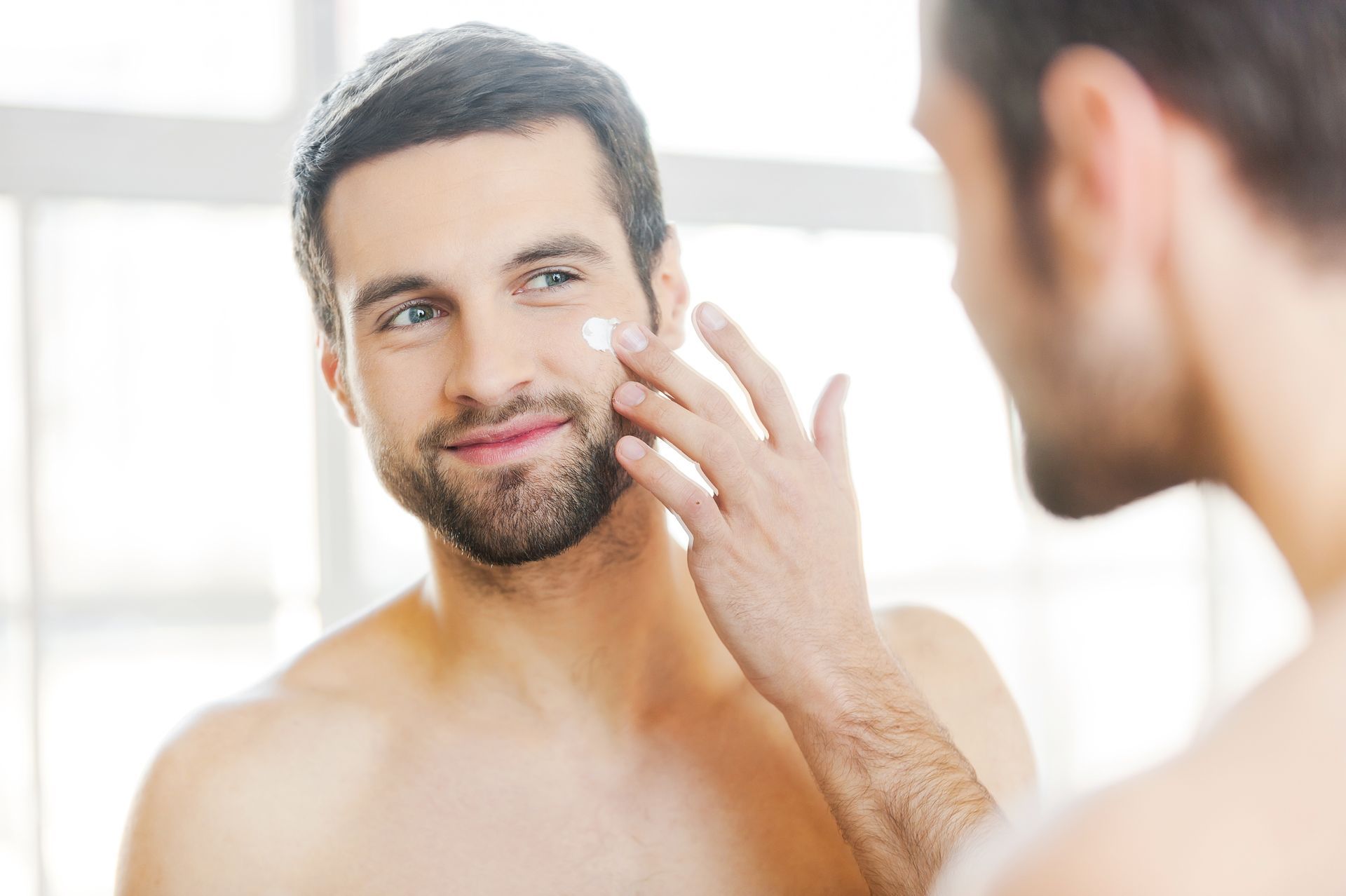 A man is applying cream to his face in front of a mirror.