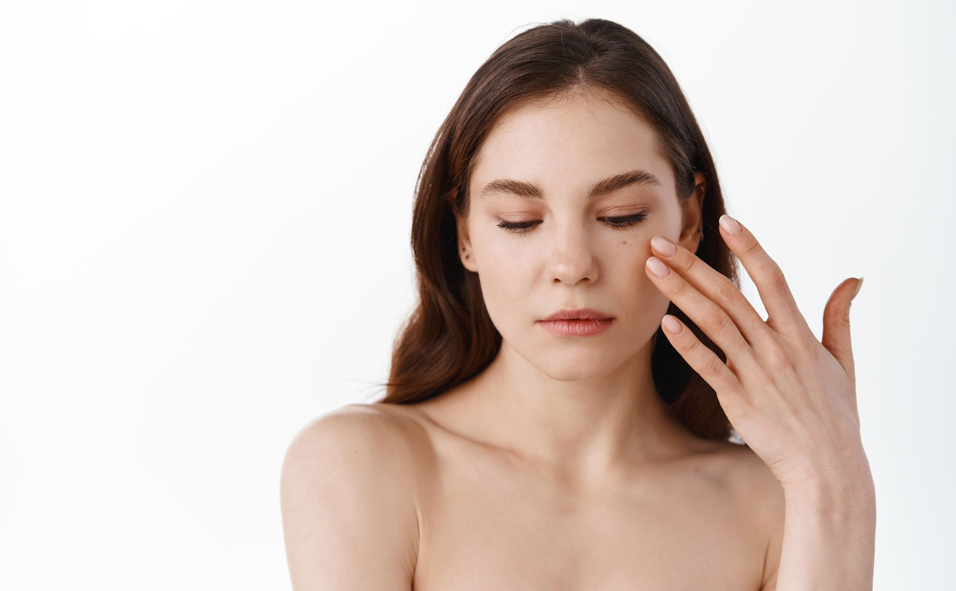 A person with long dark hair looking down while gently touching their cheek against a plain white background.