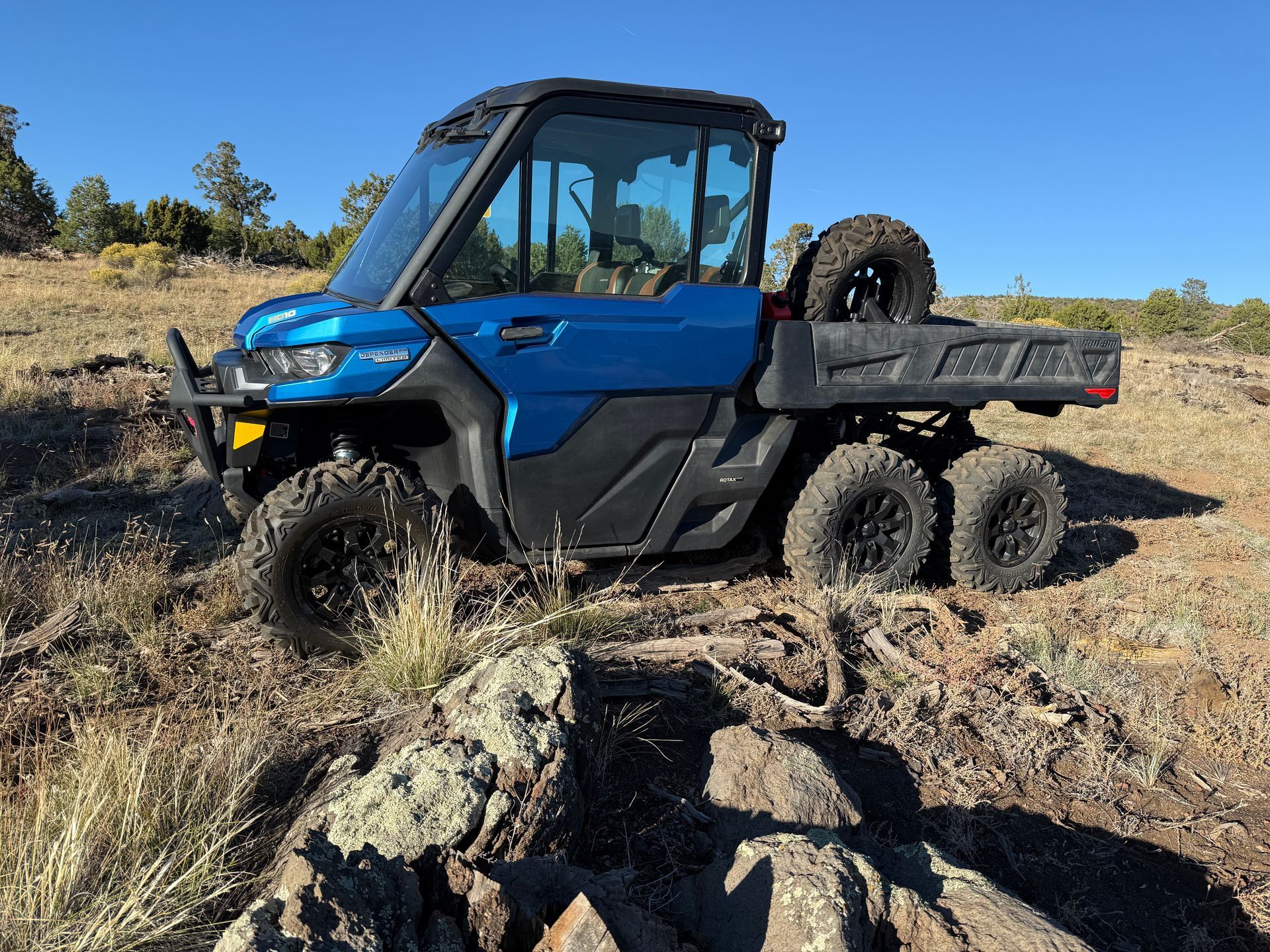 Blue 6x6 Can-Am Defender UTV parked on a rocky, grassy hillside under a clear, sunny sky.