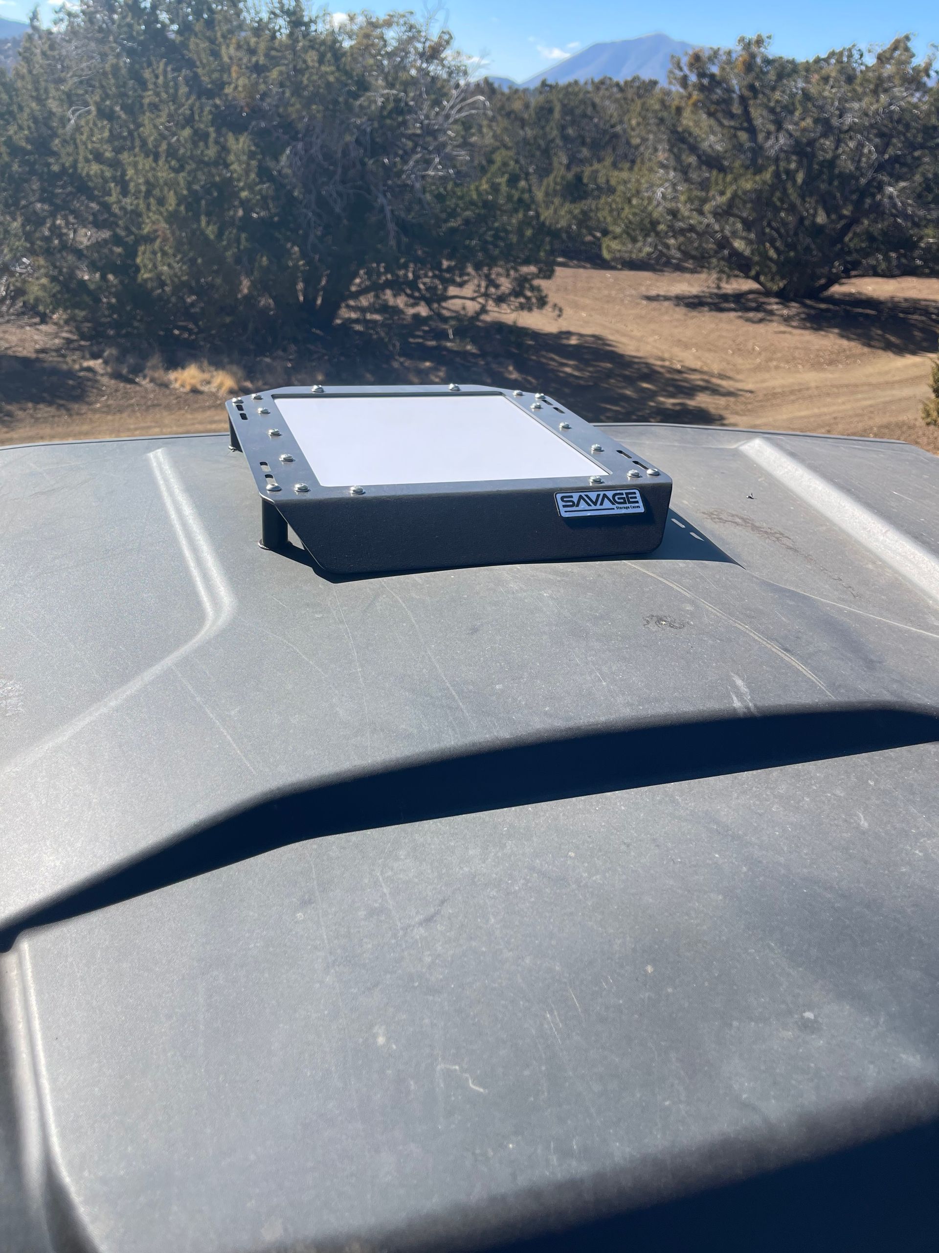 Black, rectangular device with a screen on a dark surface, outdoors with trees and mountains.