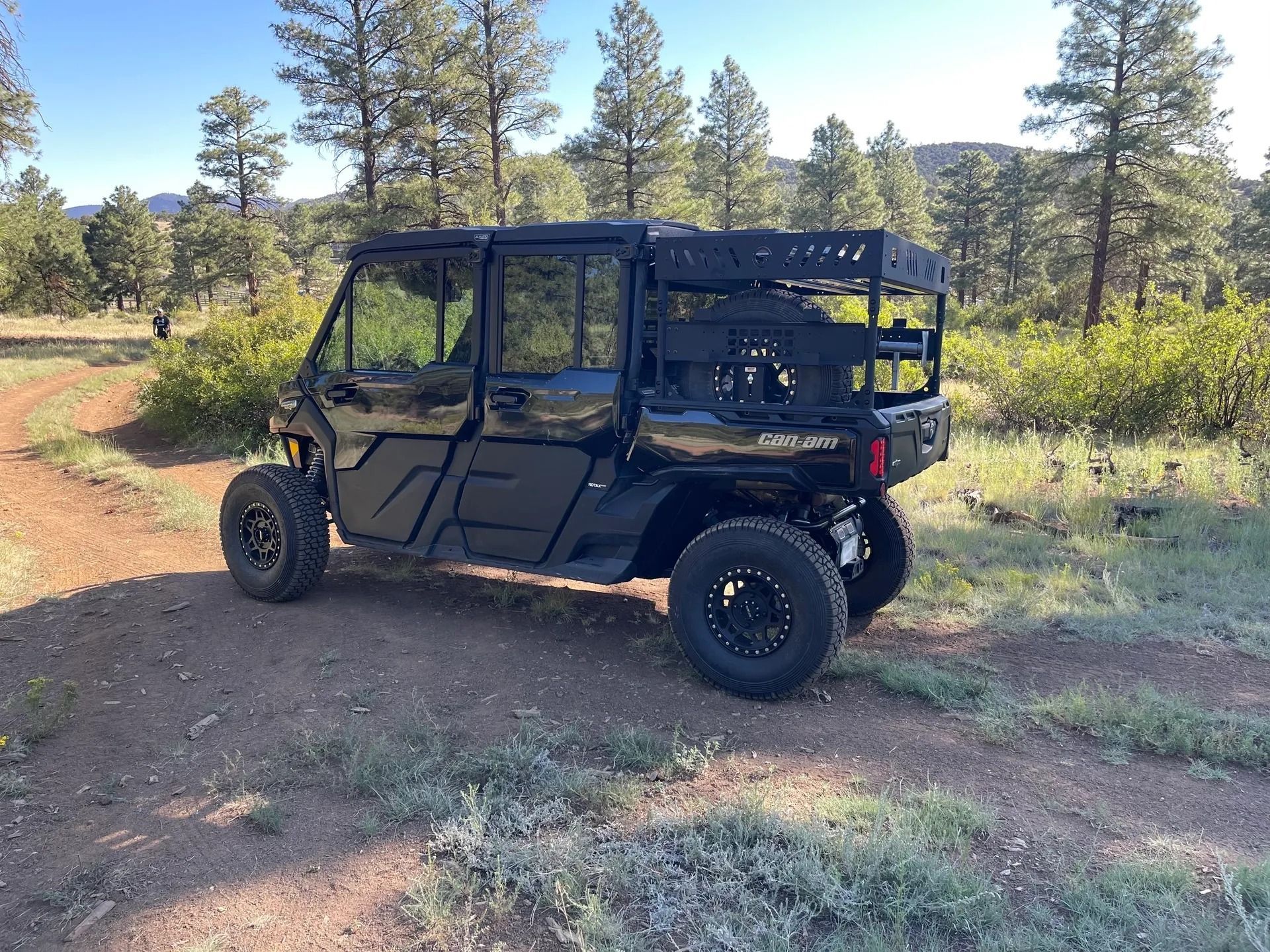 Black off-road vehicle with roof rack parked on a dirt trail, surrounded by trees.