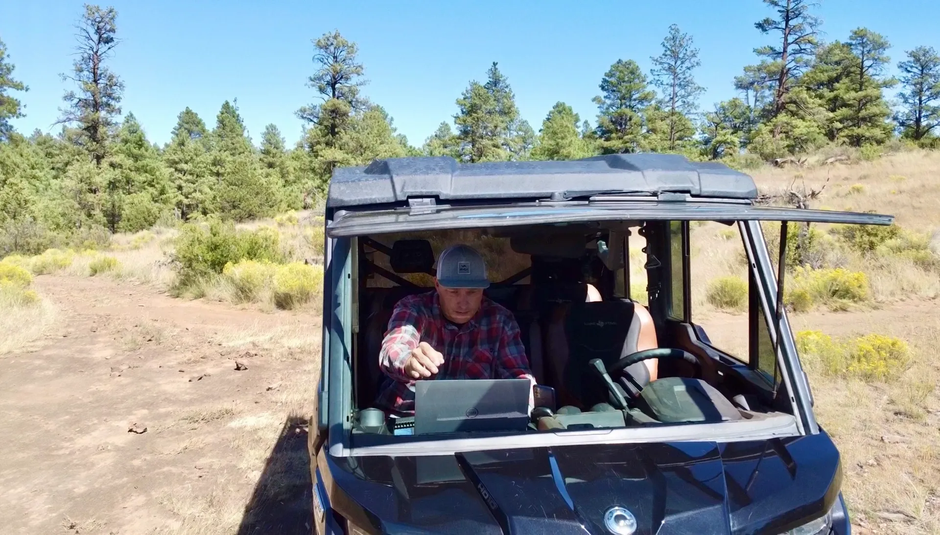 Man in a vehicle with a laptop, gesturing outdoors, surrounded by trees and a dirt path.