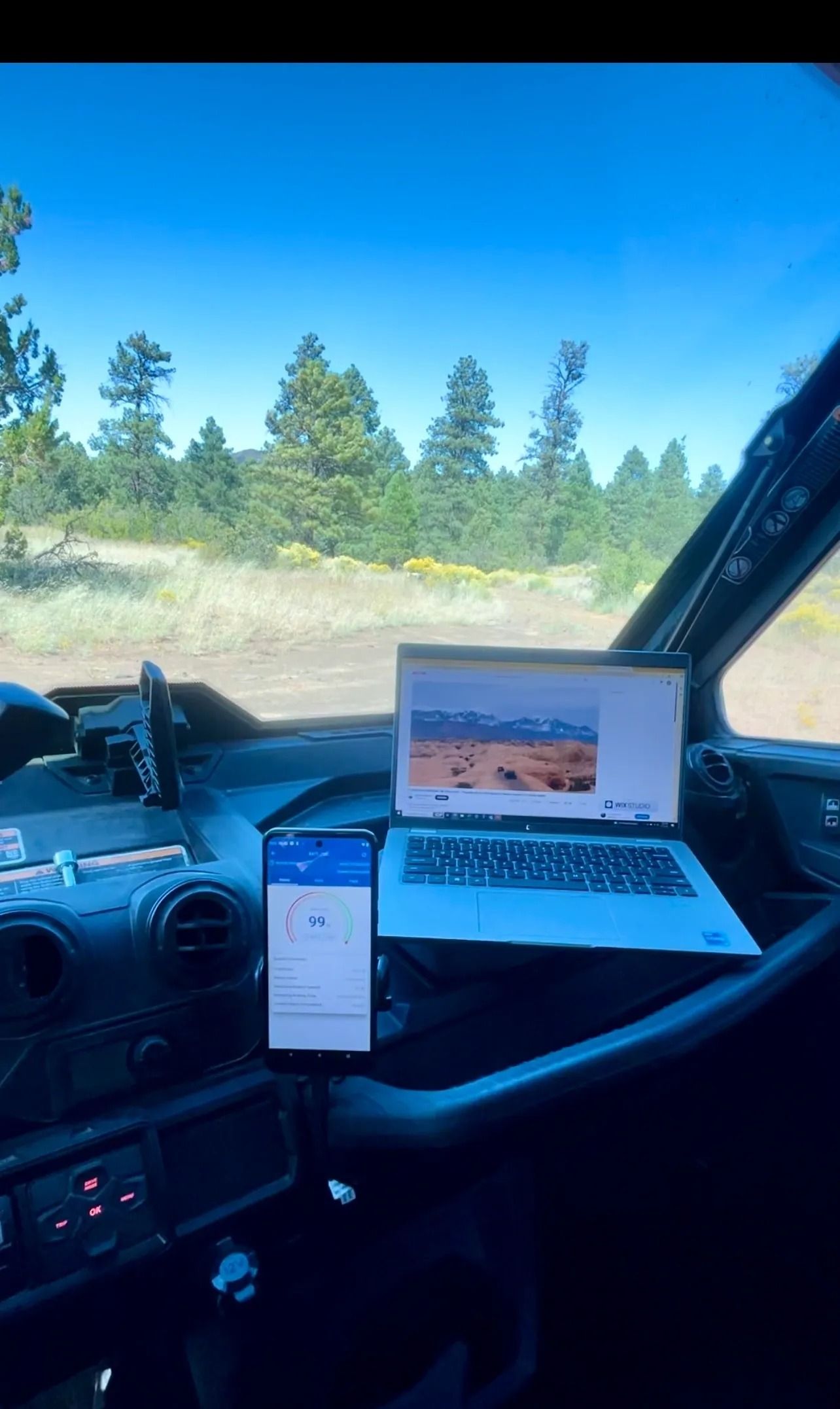 Dashboard view: Laptop and phone display maps in a vehicle. Forest visible outside.