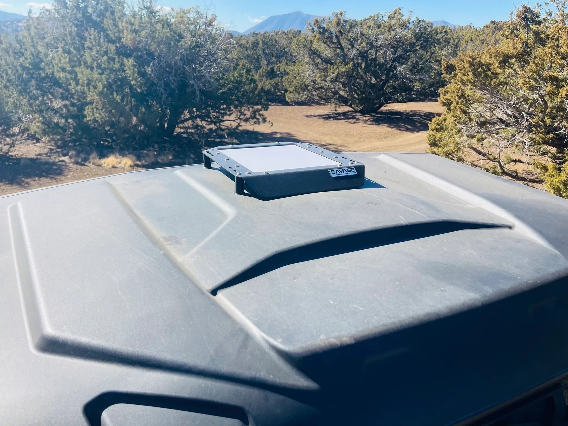 Black vehicle hood with a square, dark-colored solar panel, surrounded by trees and mountains in the background.