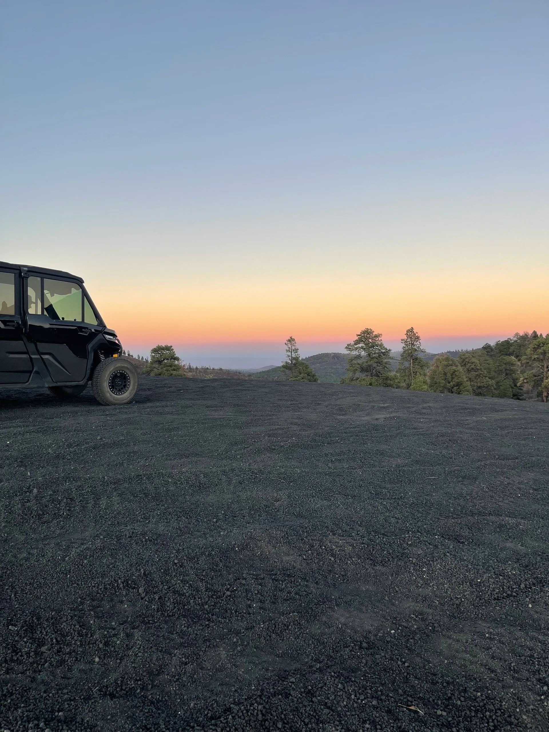 Black UTV on a gravel hill at sunset. Orange and blue sky over distant trees.