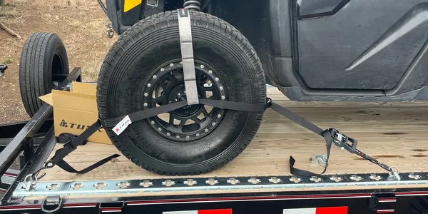 A black ATV tire is secured to a trailer with straps. A cardboard box sits nearby.