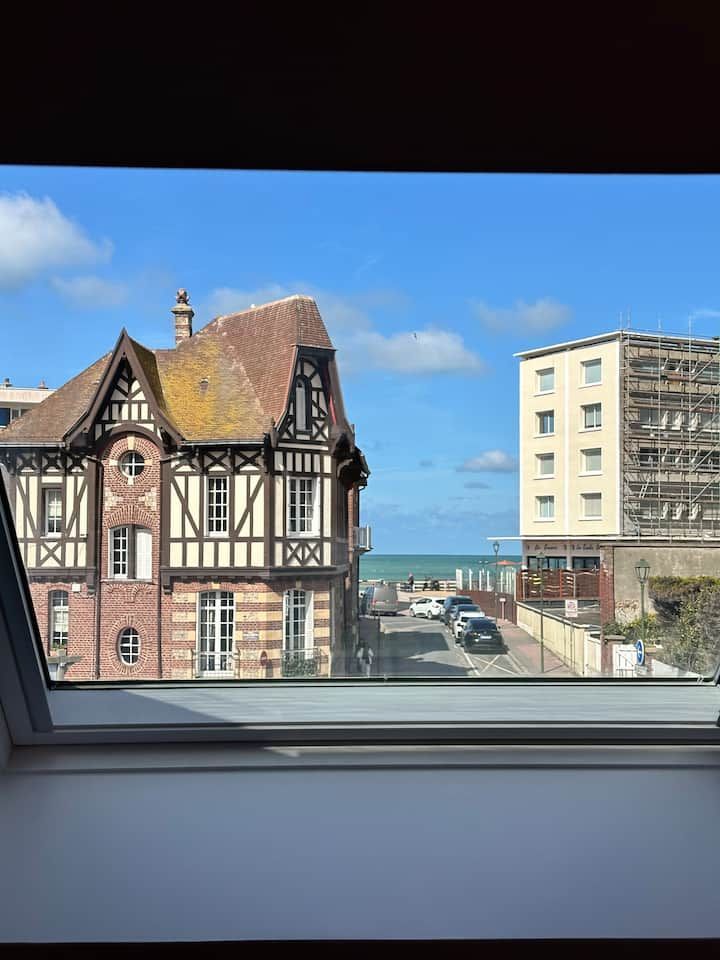 Vue par une fenêtre sur une rue en bord de mer, un immeuble à colombages, un immeuble d'appartements et un ciel bleu.