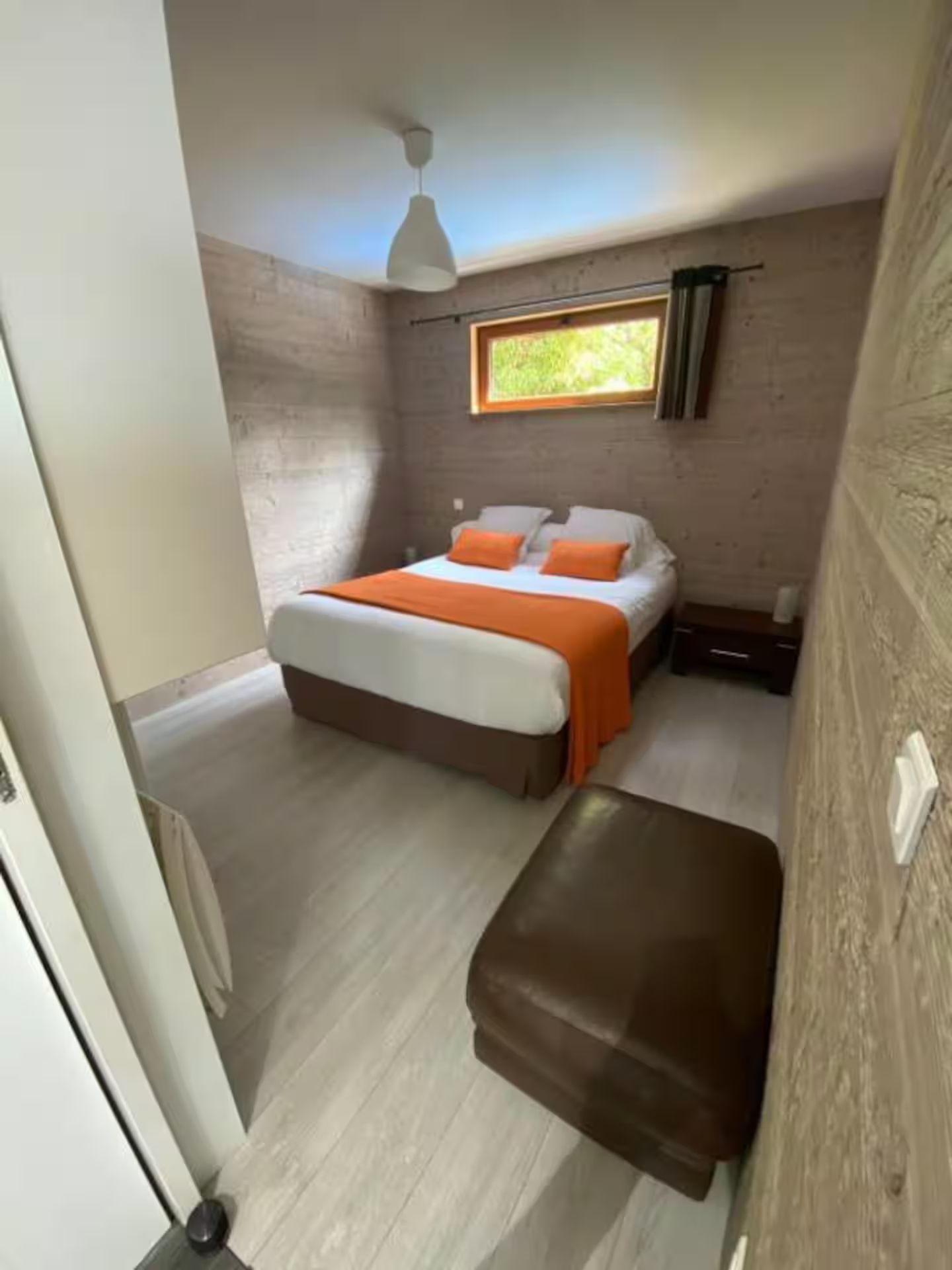 Une chambre avec un lit, un pouf, une table de chevet et une fenêtre.