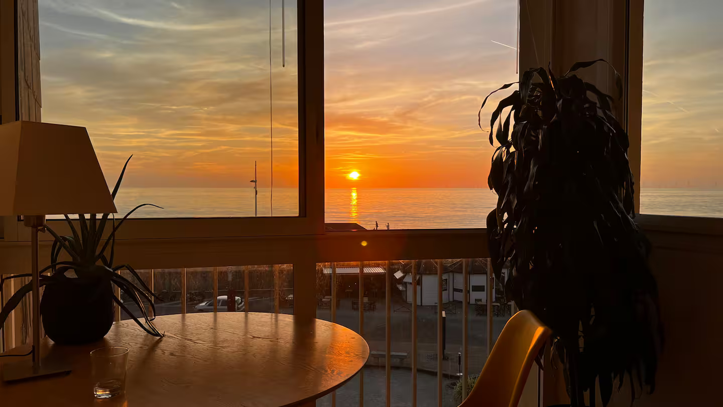 Un coucher de soleil sur l'océan est visible à travers une fenêtre dans un salon.